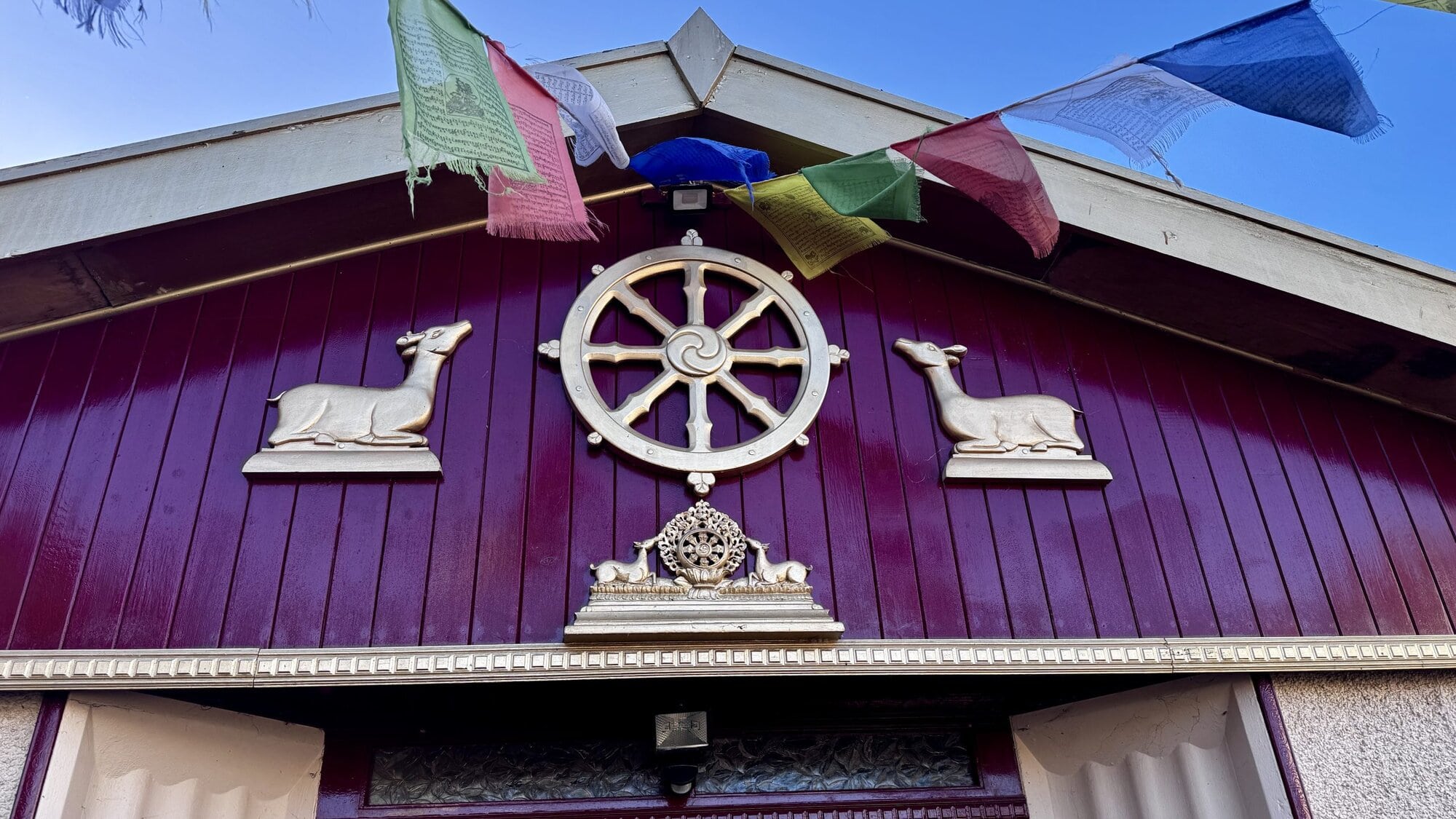 A close-up shot of the maroon entrance gable of the Sakya Thubten Ling Buddhist centre, showing a golden Dharma wheel flanked by two deer, with colourful Tibetan prayer flags fluttering against a blue sky.