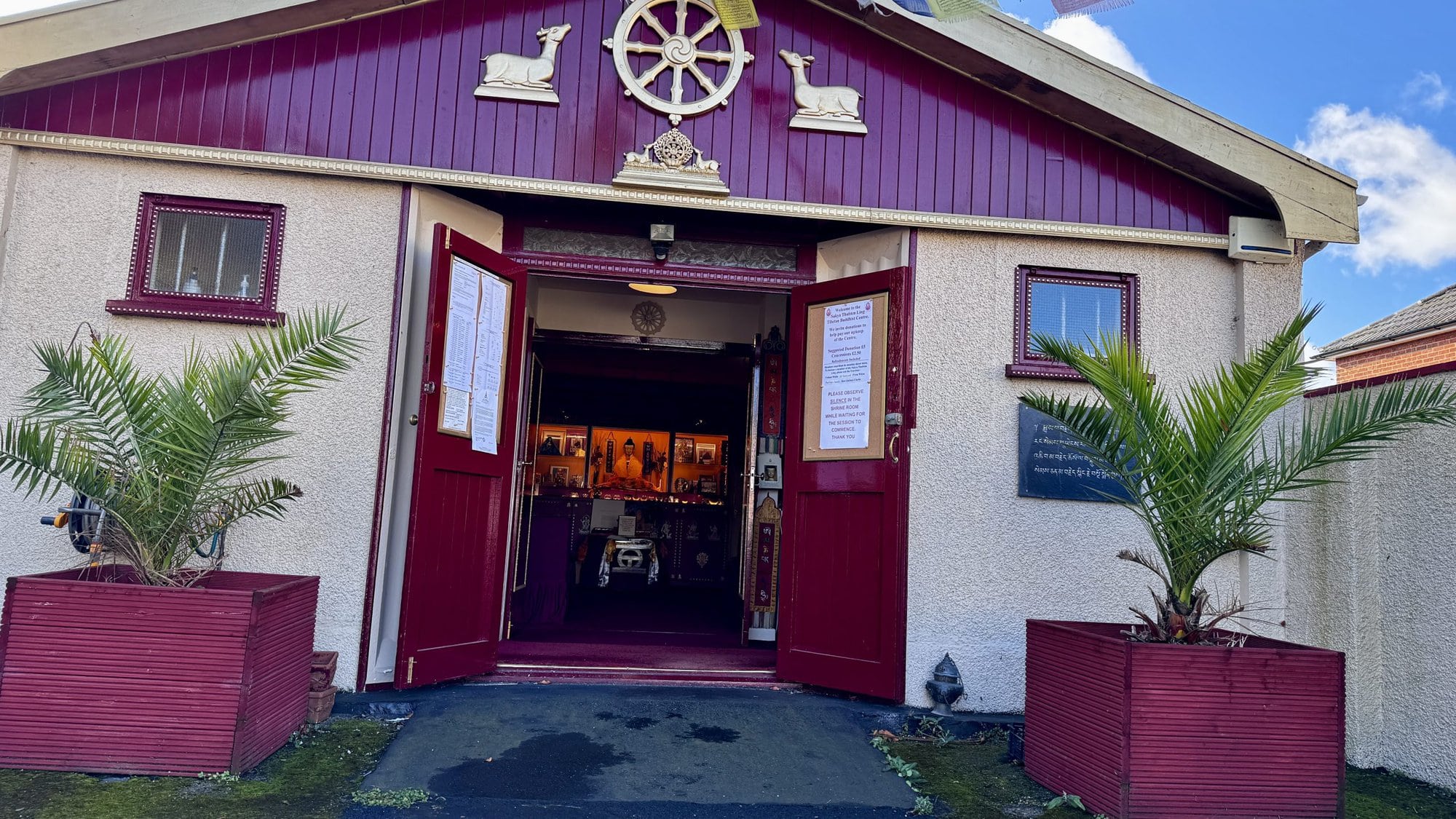 The open maroon doors of the Sakya Thubten Ling Buddhist Centre in Bournemouth, showing a view into the shrine room, with a golden Dharma wheel and prayer flags above the entrance.