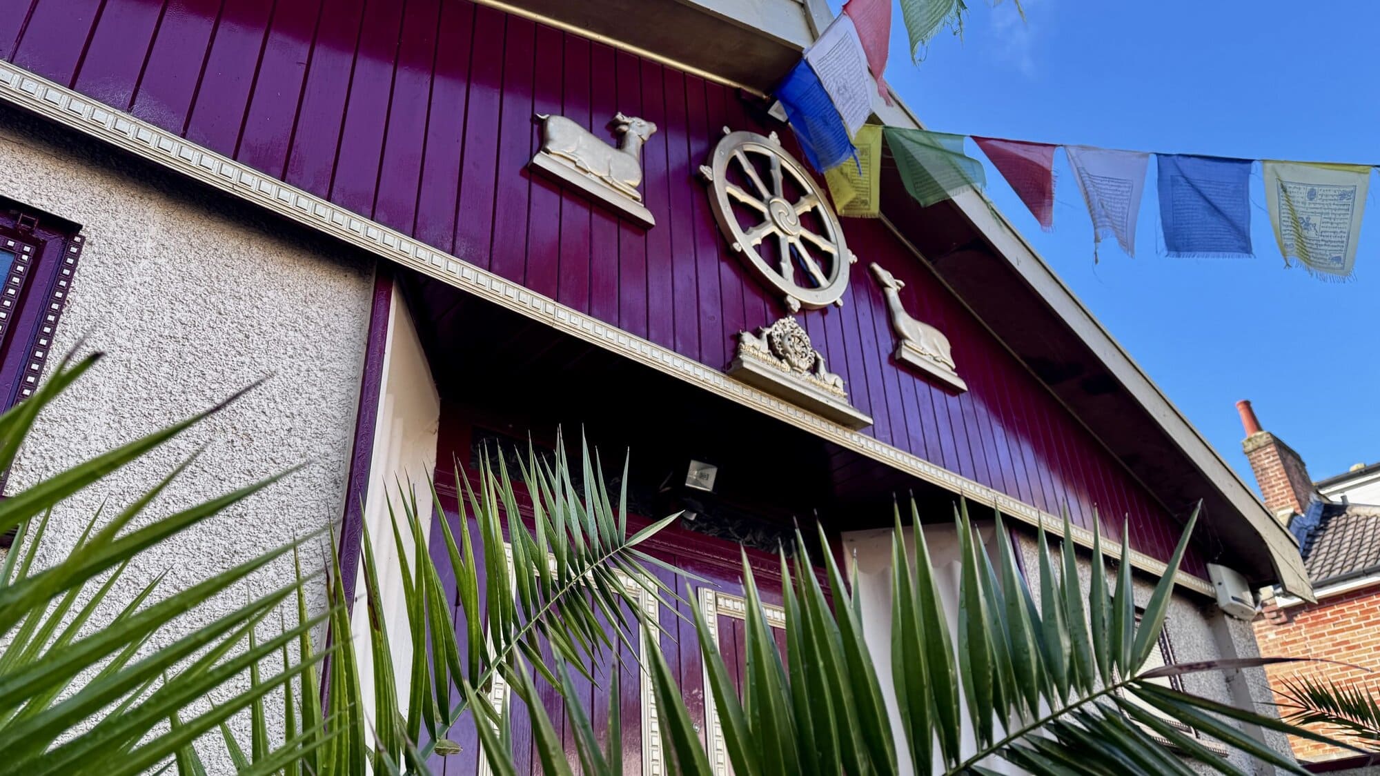 A close-up of the maroon entrance gable of Sakya Thubten Ling, showing the golden Dharma wheel flanked by two deer, with colourful Tibetan prayer flags against a blue sky.